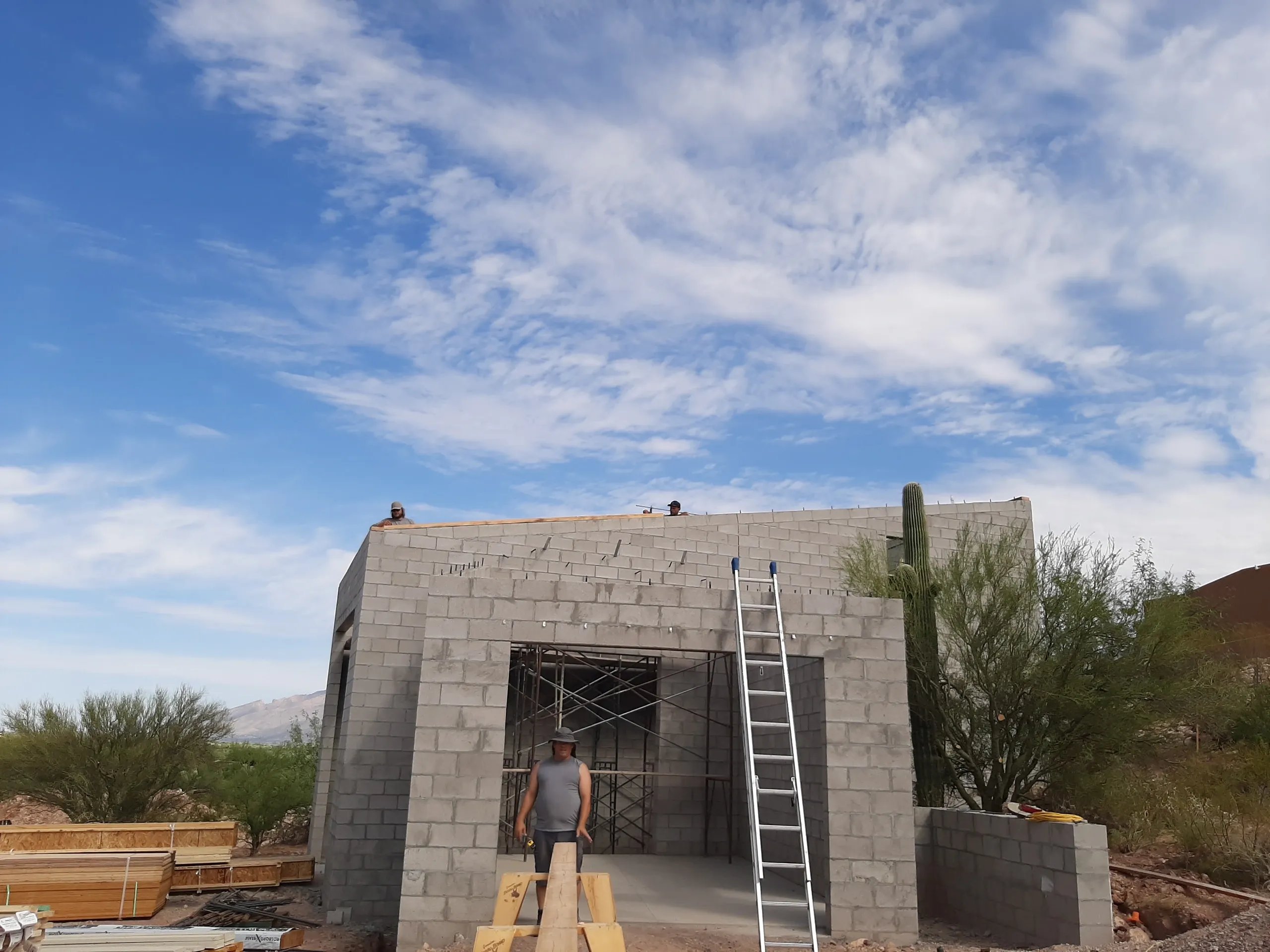 Concrete block building under construction with ladder, workers, and desert landscape